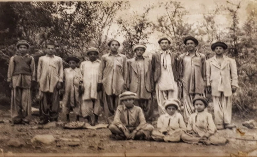 Black and white photo of men and boys in a rural village outdoors.