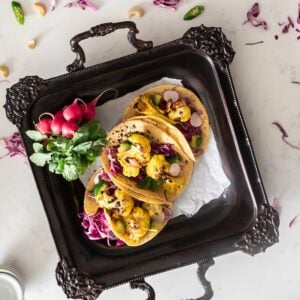 Tray with three corn cauliflower tacos with purple cabbage and avocado cubes