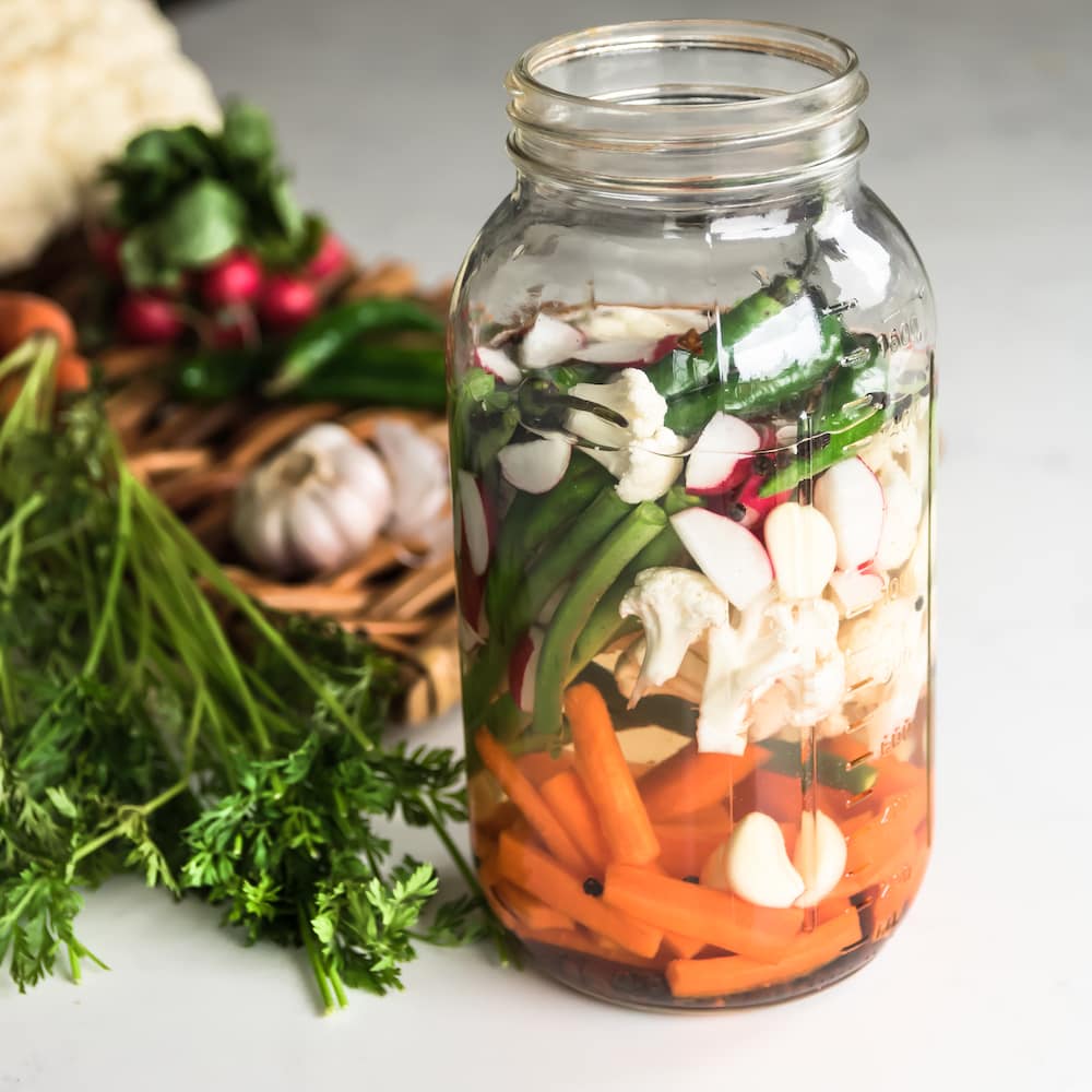 jar of mixed vegetables submerged in water with vegetables on a tray in the background.