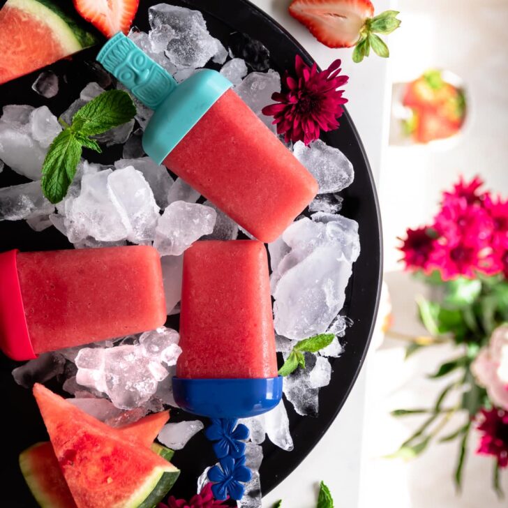 3 strawberry popsicles on ice on a round plate decoratd with fresh watermelon and flowers