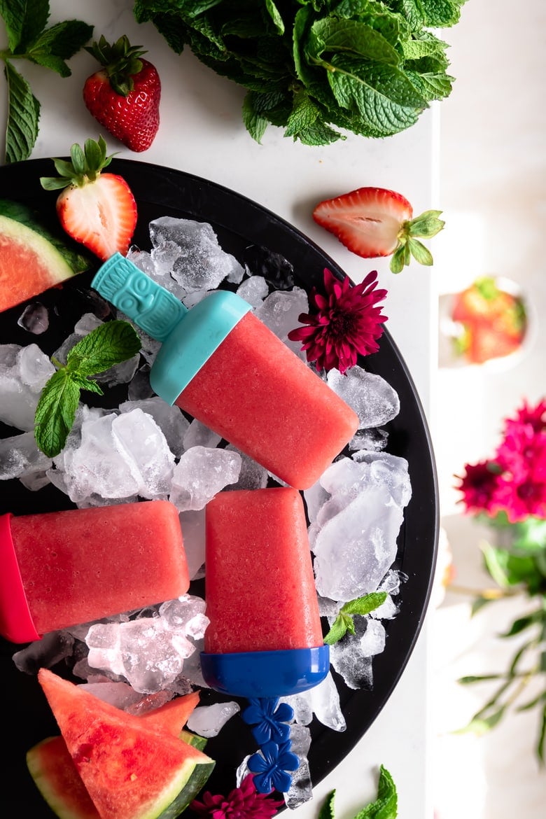 3 strawberry popsicles on ice on a round plate decoratd with fresh watermelon and flowers