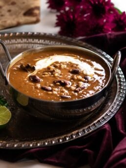daal makhani in a silver bowl on a round tray on top of a maroon velvet cloth with maroon flowers and roti in the background