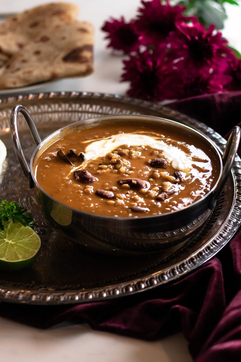 daal makhani in a silver bowl on a round tray on top of a maroon velvet cloth with maroon flowers and roti in the background