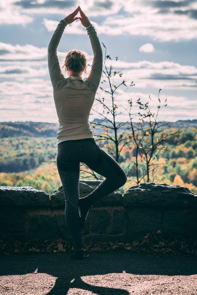 a woman doing the tree yoga pose outdoors, overlooking greenery