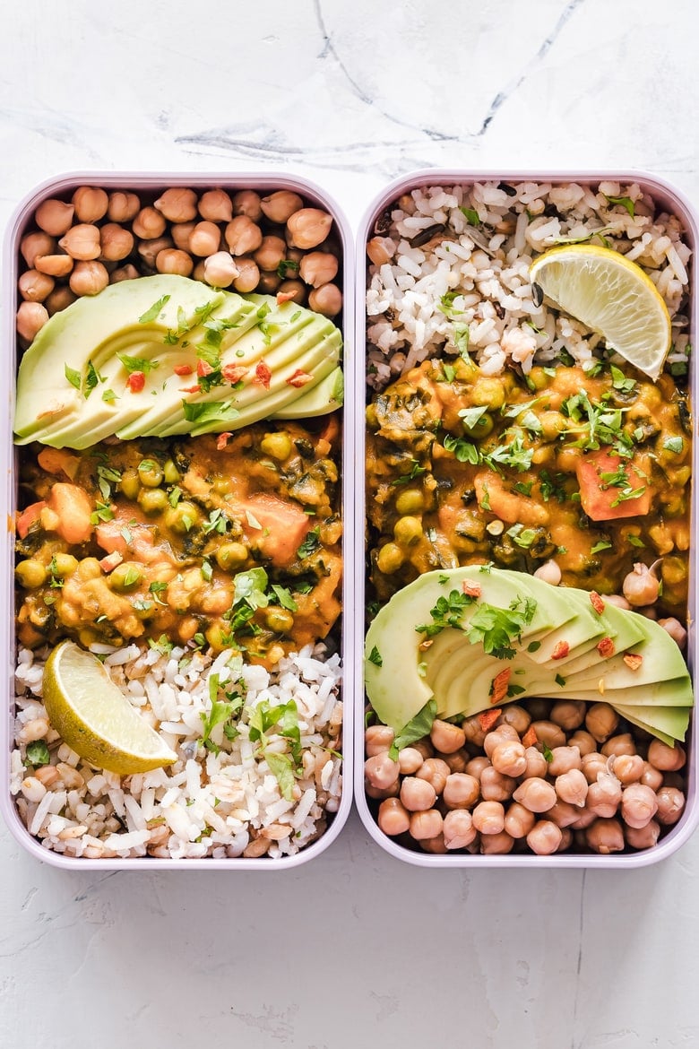 two rectangular lunch boxes with chickpeas, curry, rice and sliced avocados