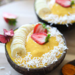 two coconut bowls filled with pineapple mango smoothie decorated with sliced bananas and fresh strawberries