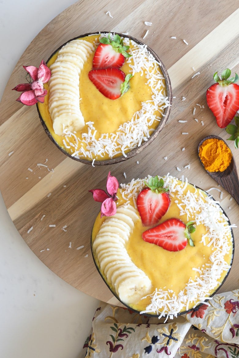 two coconut bowls filled with pineapple mango smoothie decorated with sliced bananas and fresh strawberries