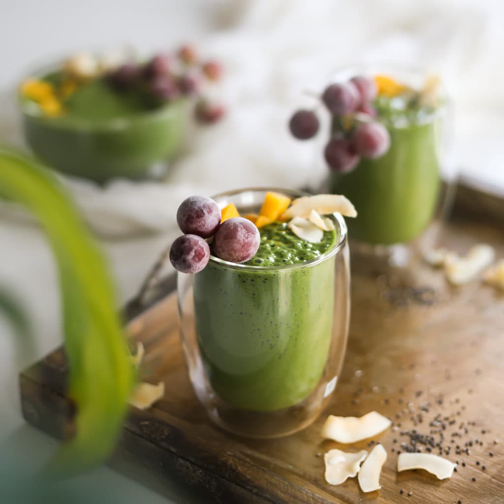 green smoothie glasses in a tray topped with frozen grapes, mango segments and coconuts flakes