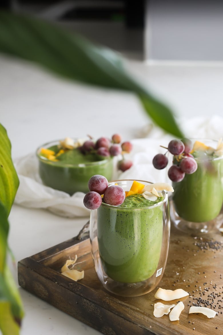 green smoothie glasses in a tray topped with frozen grapes, mango segments and coconuts flakes