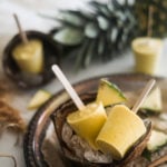 Pineapple Coconut Ice Lollies in a coconut shell on a bed of ice in a tray with a spoon of turmeric and sliced pineapples around