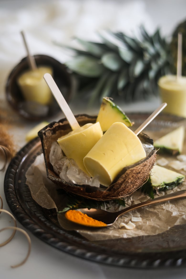 Pineapple Coconut Ice Lollies in a coconut shell on a bed of ice in a tray with a spoon of turmeric and sliced pineapples around