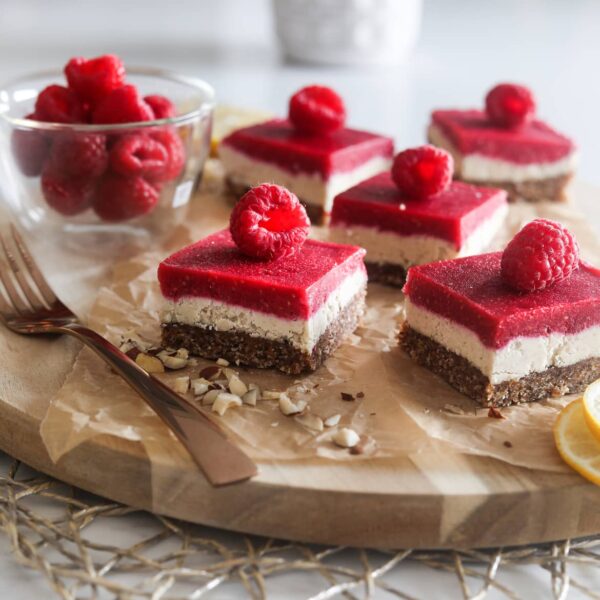 five raspberry vegan bars with a raspberry on each slice, placed on a wooden board with sprinkle of chopped nuts and a gold fork