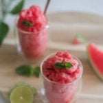 two glasses of watermelon slushie surrounded by pieces of lime and watermelon