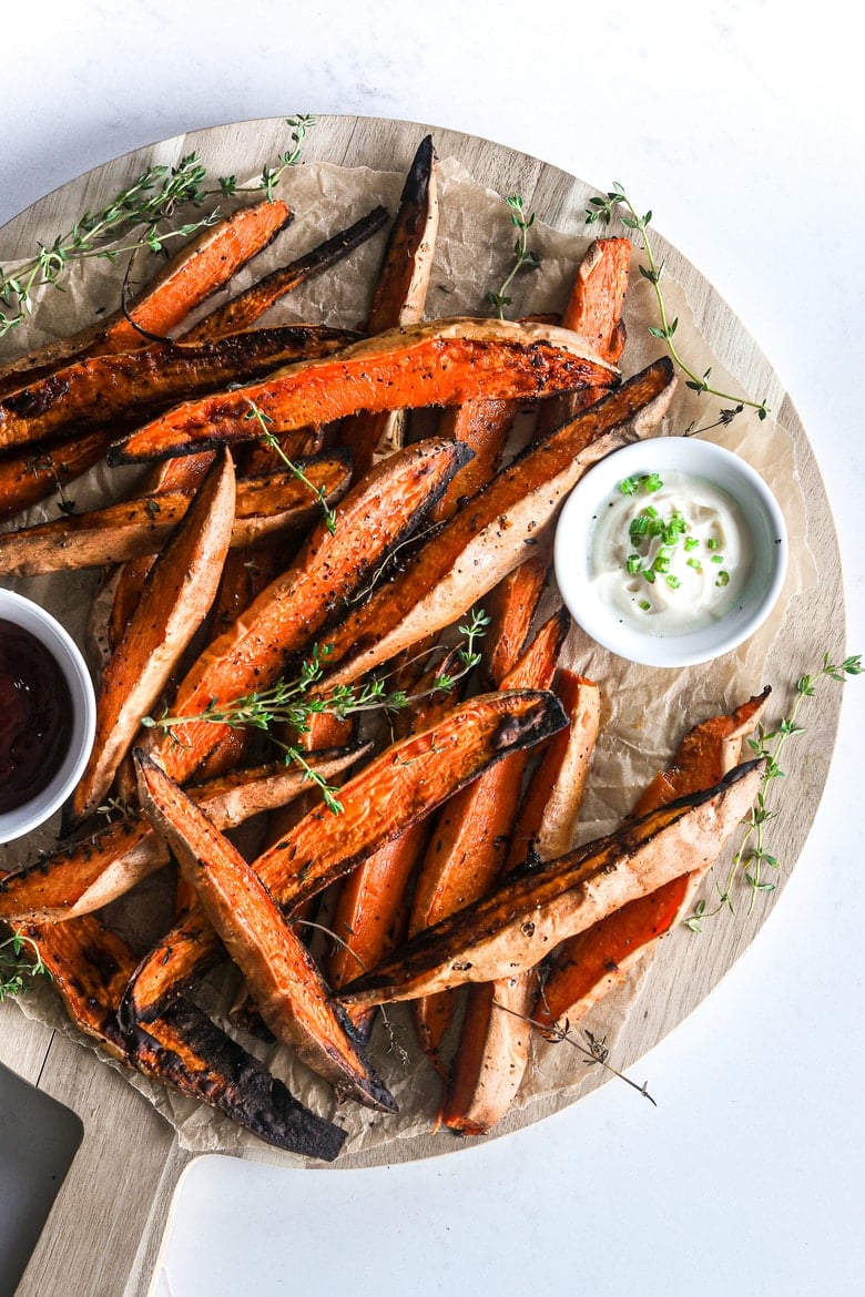 pile of baked sweet potato wedges on a wooden round board with two bowls of dipping sauces