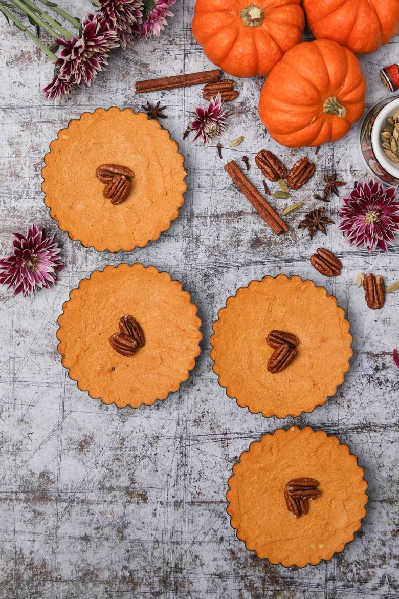 a row of homemade pumpkin pies topped with pecans with pumpkins and flowers in the background