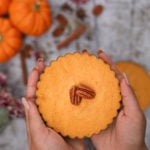 hands holding a homemade pumpkin pies topped with pecans with pumpkins and spices in the background