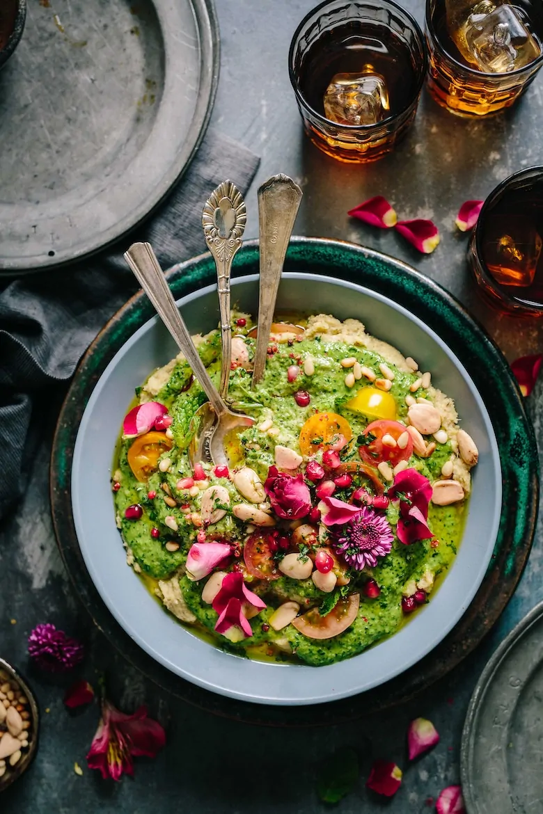 Bowl of soup topped with green chutney , vegetables and flowers with 2 glasses filled with and empty plates around