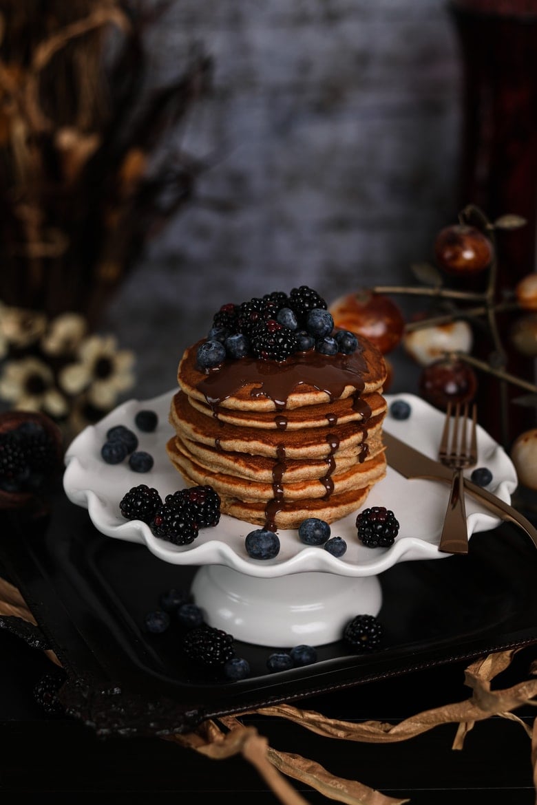 stack of fluffy pancakes on a cake stand topped with blackberries and blueberries and dark chocolate with flowers and decorations all around