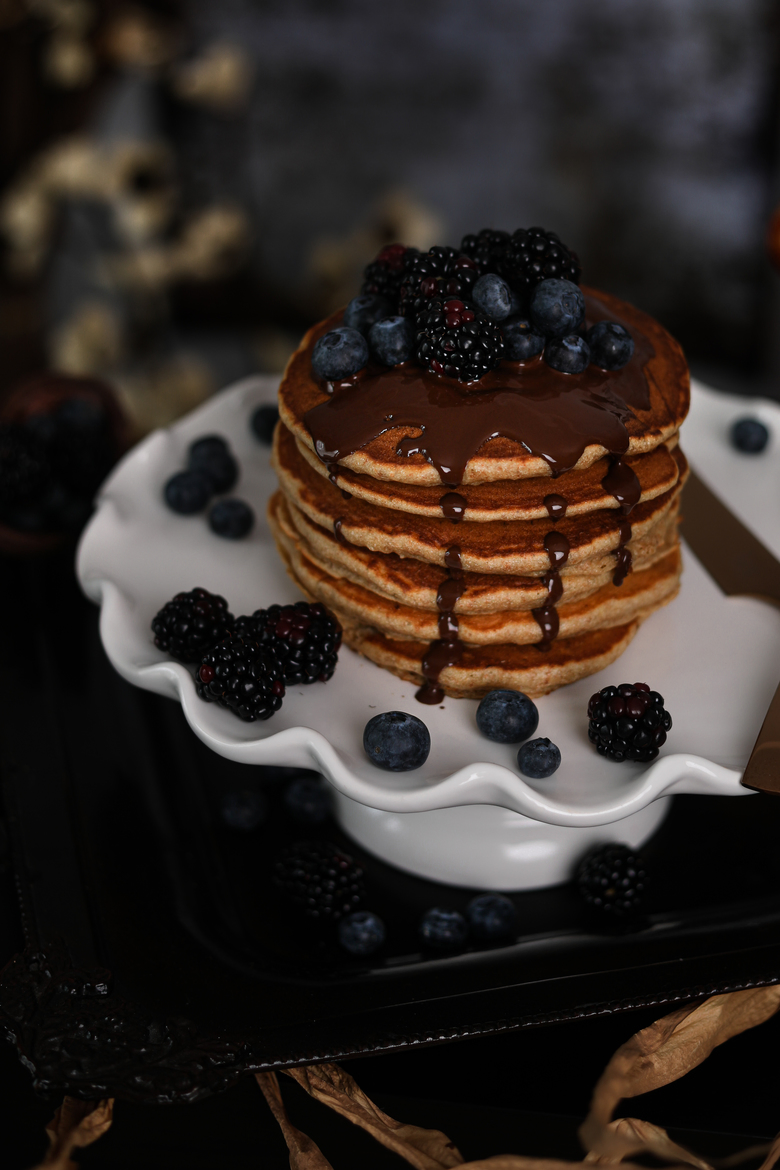 stack of fluffy pancakes on a cake stand topped with blackberries and blueberries and dark chocolate with flowers in the background