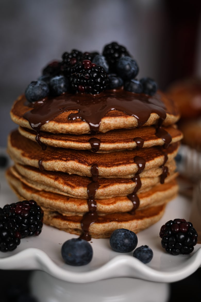 stack of fluffy pancakes on a cake stand topped with blackberries and blueberries and dark chocolate