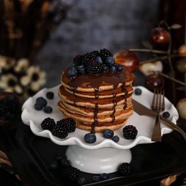 stack of fluffy pancakes on a cake stand topped with blackberries and blueberries and dark chocolate with flowers and decorations all around