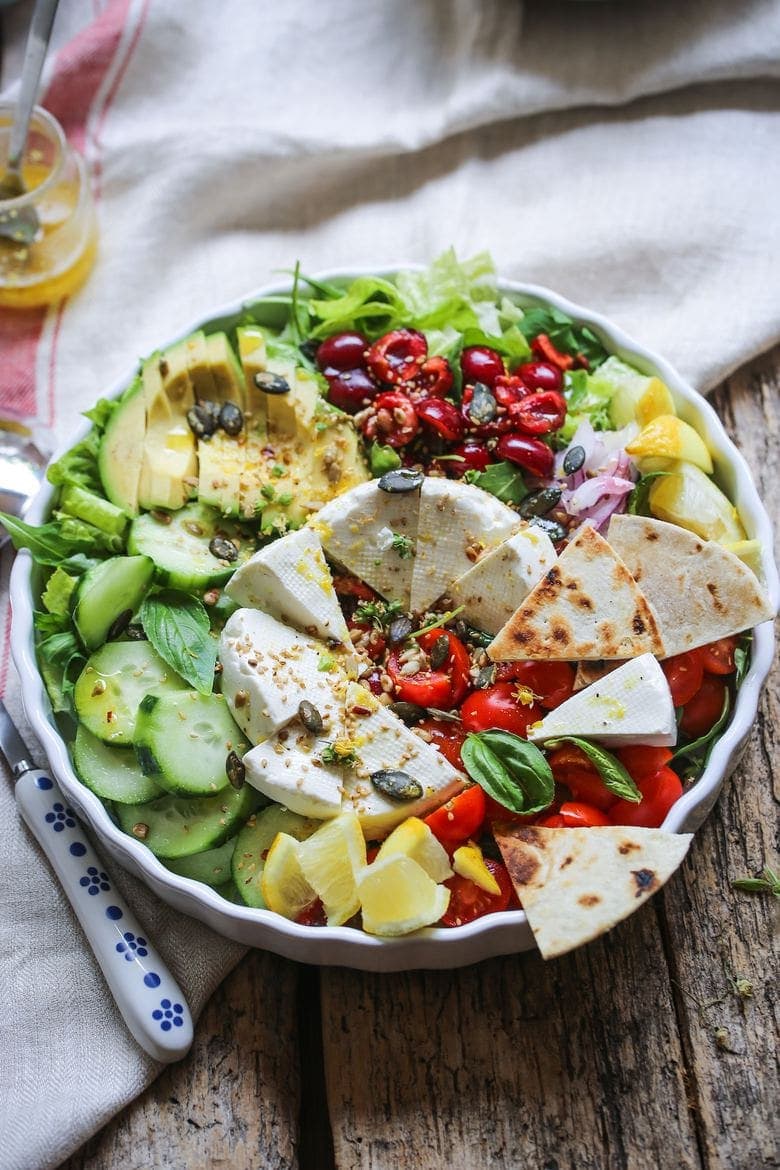 bowl full of salad topped with avocado, cucumber, tomatoes, cheese and naan bread
