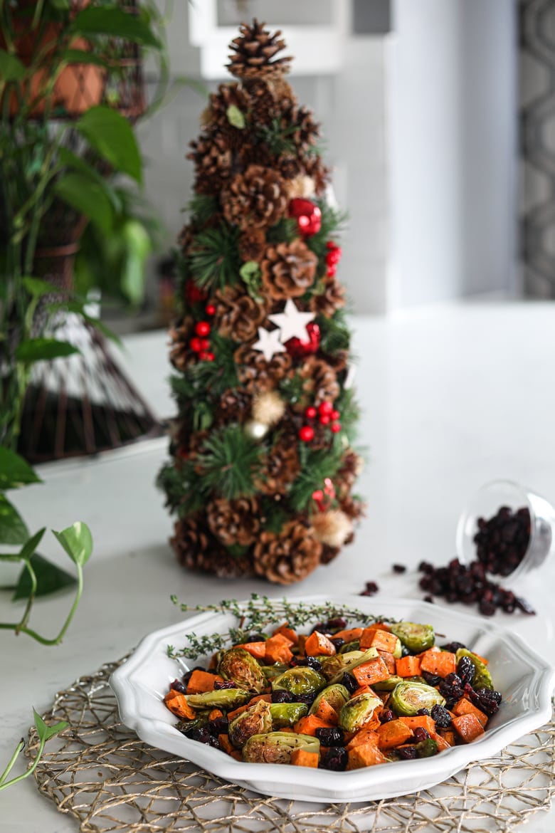 Plate of easy and healthy roasted Brussel sprouts recipe with sweet potatoes and cranberries with a Christmas tree in the background