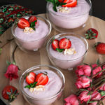 Four bowls of coconut strawberry mousse on a wooden round tray with pink roses and fresh strawberries all around