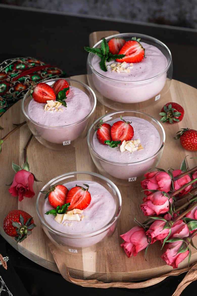 Four bowls of coconut strawberry mousse on a wooden round tray with pink roses and fresh strawberries all around