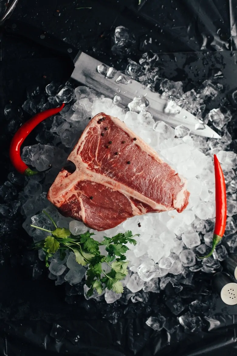 a piece of red meat on crushed ice with red chillies and a sprig of herbs next to it