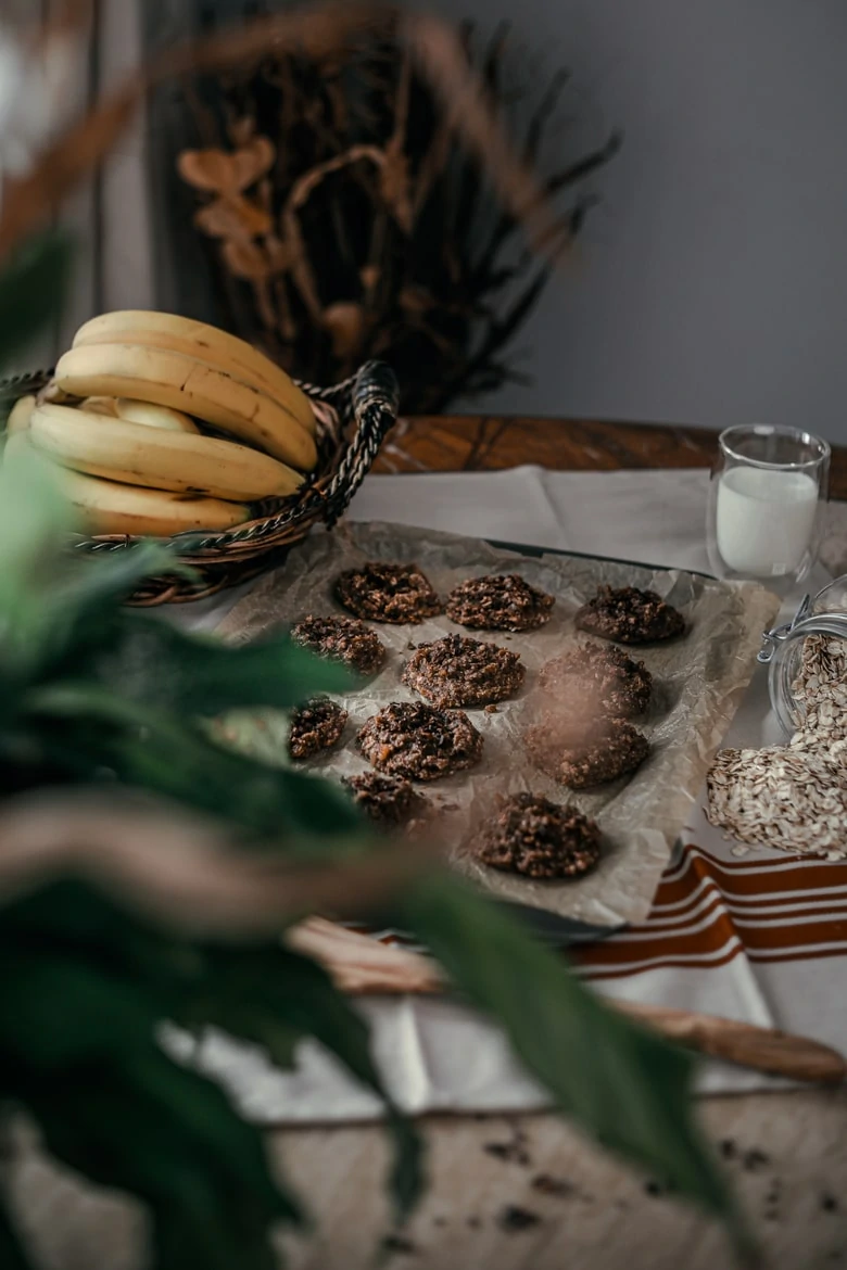 A tray of chocolate oatmeal cookies on a table with a bunch of bananas, rolled oats and a glass of milk