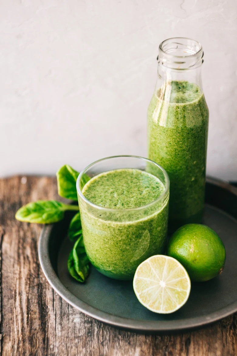 a bottle and glass of green juice in a tray with a couple of limes and spinach leaves