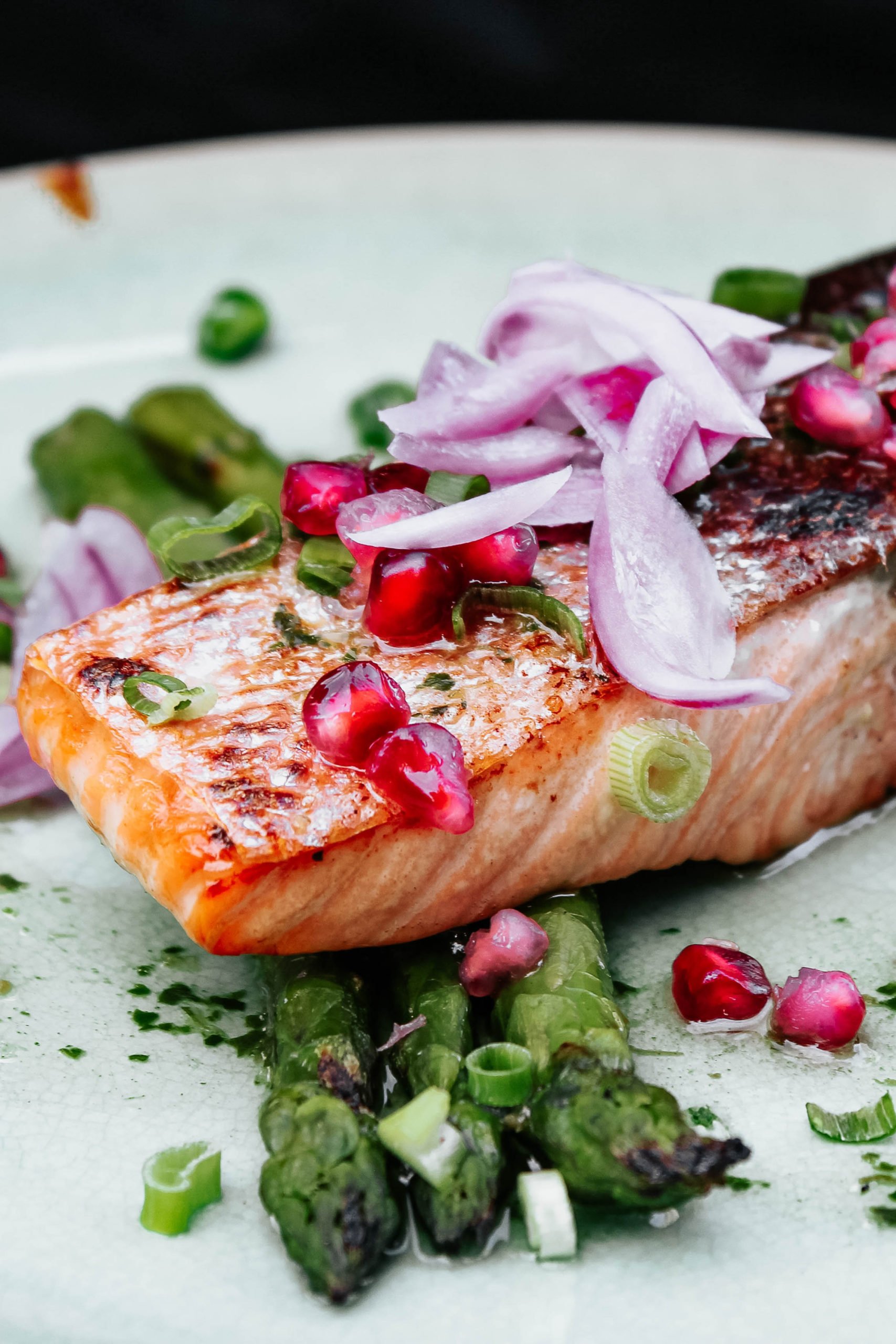 a piece of cooked salmon fillet on a bed of asparagus topped with chopped red onion and pomegranate seeds