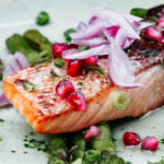 a piece of cooked salmon fillet on a bed of asparagus topped with chopped red onion and pomegranate seeds