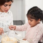 Profile view of pretty little girl with two braids adding flour into bowl while cooking appetizing pie with mom, interior of modern kitchen on background