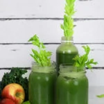 green juice in two mason jars and a bottle with celery stalks inside for decoration, surrounded by apples, kale and lime