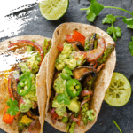 BBQ mushrooms and mixed vegetables fajitas topped with guacamole on a charcoal surface with lime wedges and herbs