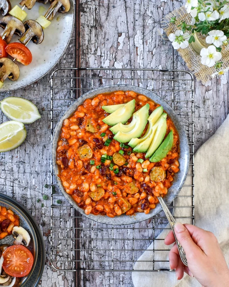A hand stirring smoky BBQ baked beans topped with avocado slices