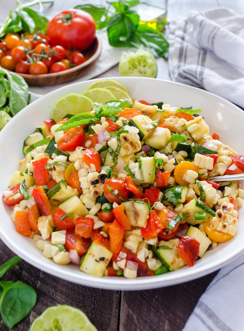 Bowl of BBQ corn and mixed vegetable salad surrounded by herbs and tomatoes