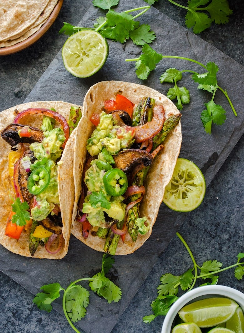 BBQ mushrooms and mixed vegetables fajitas topped with guacamole on a charcoal surface with lime wedges and herbs