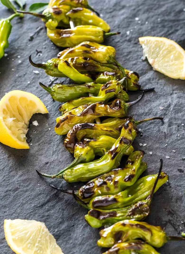 BBQ grilled shishito peppers lined up on a charcoal surface with lemon wedges