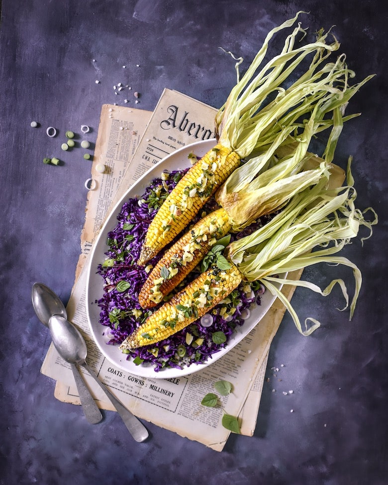 3 corn on the cob on a bed of purple cabbage placed on top of newspapers flatlay