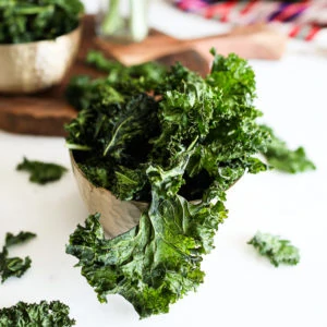 bowl of kale chips on a white work top with a board in the background with a ramekin of spice and a red shawl