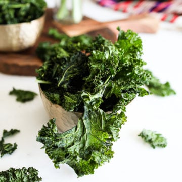 bowl of kale chips on a white work top with a board in the background with a ramekin of spice and a red shawl
