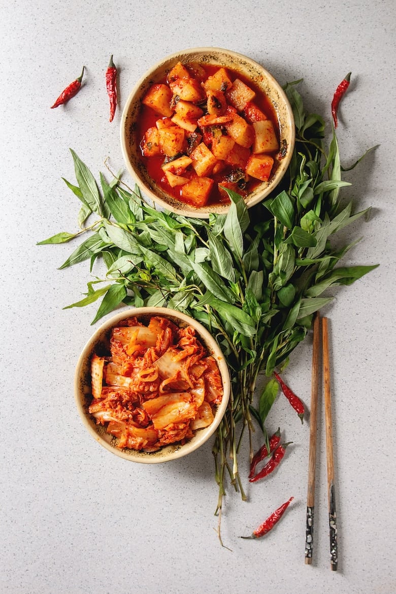 Korean traditional fermented appetizer kimchi cabbage and radish salad in ceramic plates with Vietnamese oregano greens over grey spotted background. Flat lay, space.