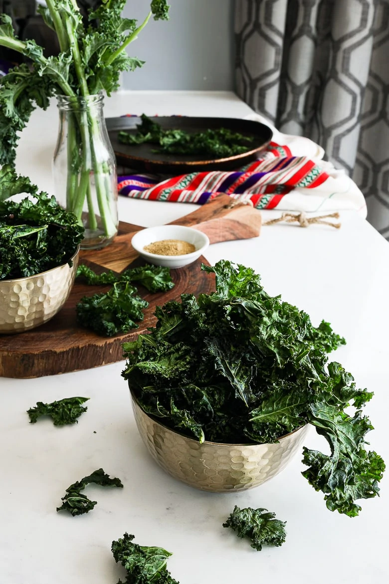 bowl of kale chips on a white work top with a board in the background with a ramekin of spice and a red shawl