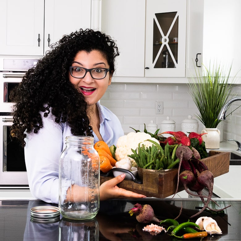 a woman holding a box of fresh vegetables standing in front of a mason jar in a kitchen with a happy shocked expression on her face.