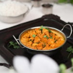 bowl of indian butter chicken on a black tray with white rice in the background and small spice containers arranged around - perspective shot