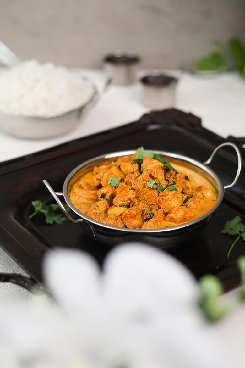 bowl of indian butter chicken on a black tray with white rice in the background and small spice containers arranged around - perspective shot