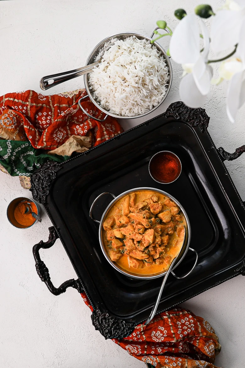 bowl of indian butter chicken on a black tray with white rice on the side and a decorative shawl - flat lay